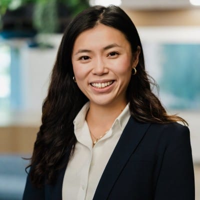 eCommerce Development 48 Professional headshot of a smiling woman with long dark hair, wearing a navy blazer over a white collared shirt against a softly blurred indoor background