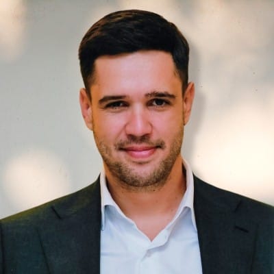 Home Page - NEW 297 Professional headshot of a smiling man with dark hair and light stubble, wearing a dark blazer over a white collared shirt against a softly blurred background