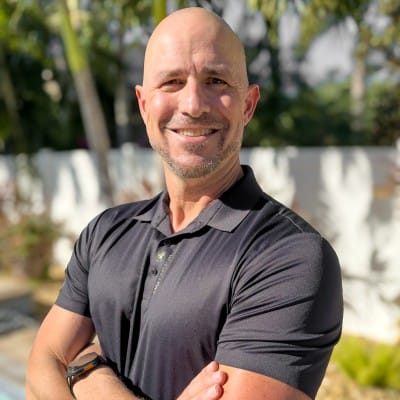 Home Page - NEW 247 Portrait of a smiling bald man with a short goatee, wearing a black polo shirt with his arms crossed outdoors in a sunny garden setting