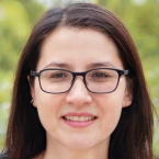 Close-up portrait of a woman with long dark hair and glasses, smiling slightly against a softly blurred green outdoor background