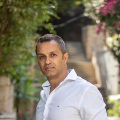 Web Design 81 Portrait of a man with short dark hair, wearing a white button-down shirt and standing outdoors in a leafy garden setting with stairs in the background