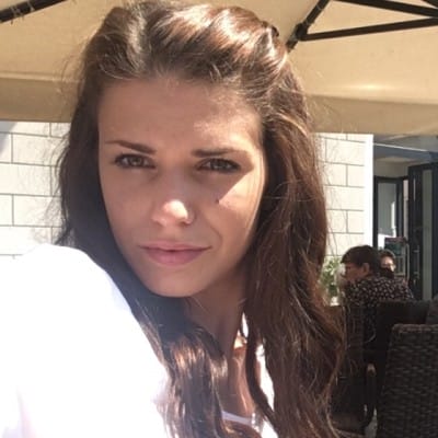 eCommerce Development 56 Close-up selfie of a woman with long brown hair, wearing a white top and looking at the camera while seated outdoors at a café in bright sunlight