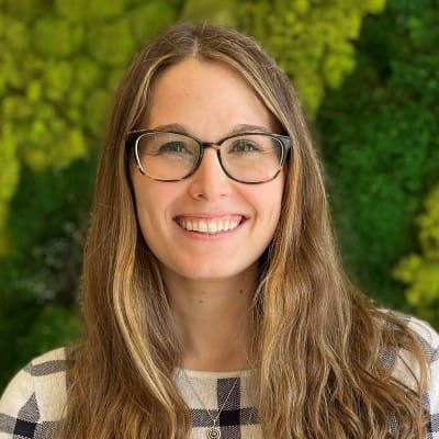 Home Page - NEW 247 Close-up portrait of a smiling woman with long light brown hair and glasses, wearing a checkered top against a leafy green outdoor background