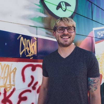 eCommerce Development 81 Smiling man with glasses and a tattooed arm wearing a dark T-shirt, standing in front of a colorful graffiti-covered wall