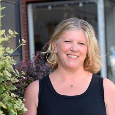 Website Redesign 71 Smiling woman with blonde hair wearing hoop earrings and a black sleeveless top, standing outdoors near plants and a storefront