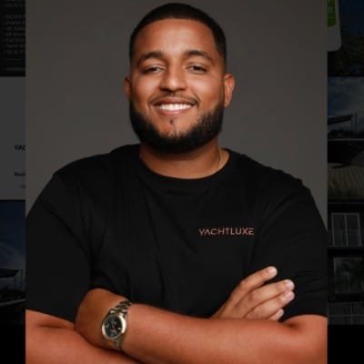 Home Page - NEW 247 Smiling man wearing a black T-shirt and wristwatch, posing with arms crossed against a dark background