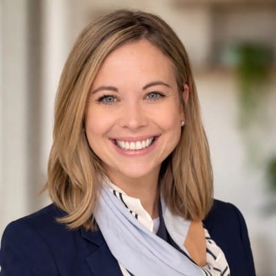 Law Firm Web Design 57 Professional headshot of a smiling woman with shoulder-length blonde hair, wearing a navy blazer and light scarf against a softly blurred indoor background