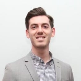 eCommerce Development 80 Portrait of a smiling man with short brown hair, wearing a light gray blazer over a checkered button-down shirt against a plain light background