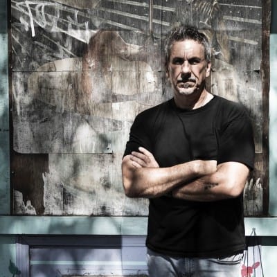 Home Page - NEW 297 Man wearing a black T-shirt and jeans, standing with arms crossed in front of a textured wall with abstract artwork