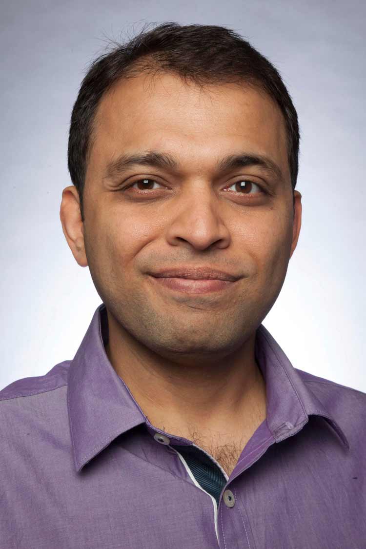 WooCommerce Development 49 Close-up professional headshot of a smiling man with short dark hair, wearing a purple collared shirt against a light gray studio background