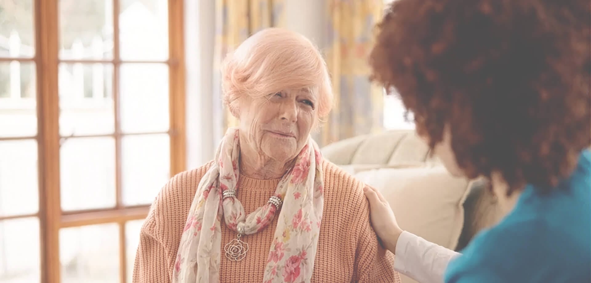 Older woman wearing a floral scarf sits indoors while another person places a comforting hand on her shoulder