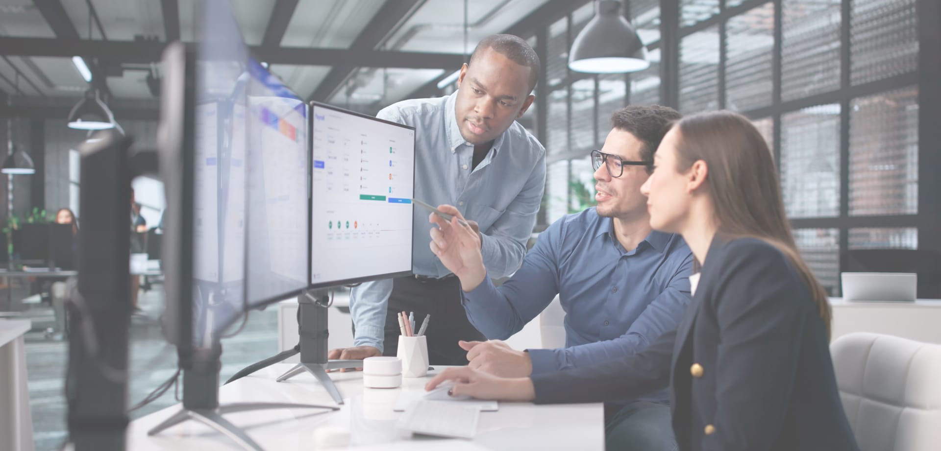 Three colleagues collaborating at desktop monitors in a modern office, reviewing information together on screen.